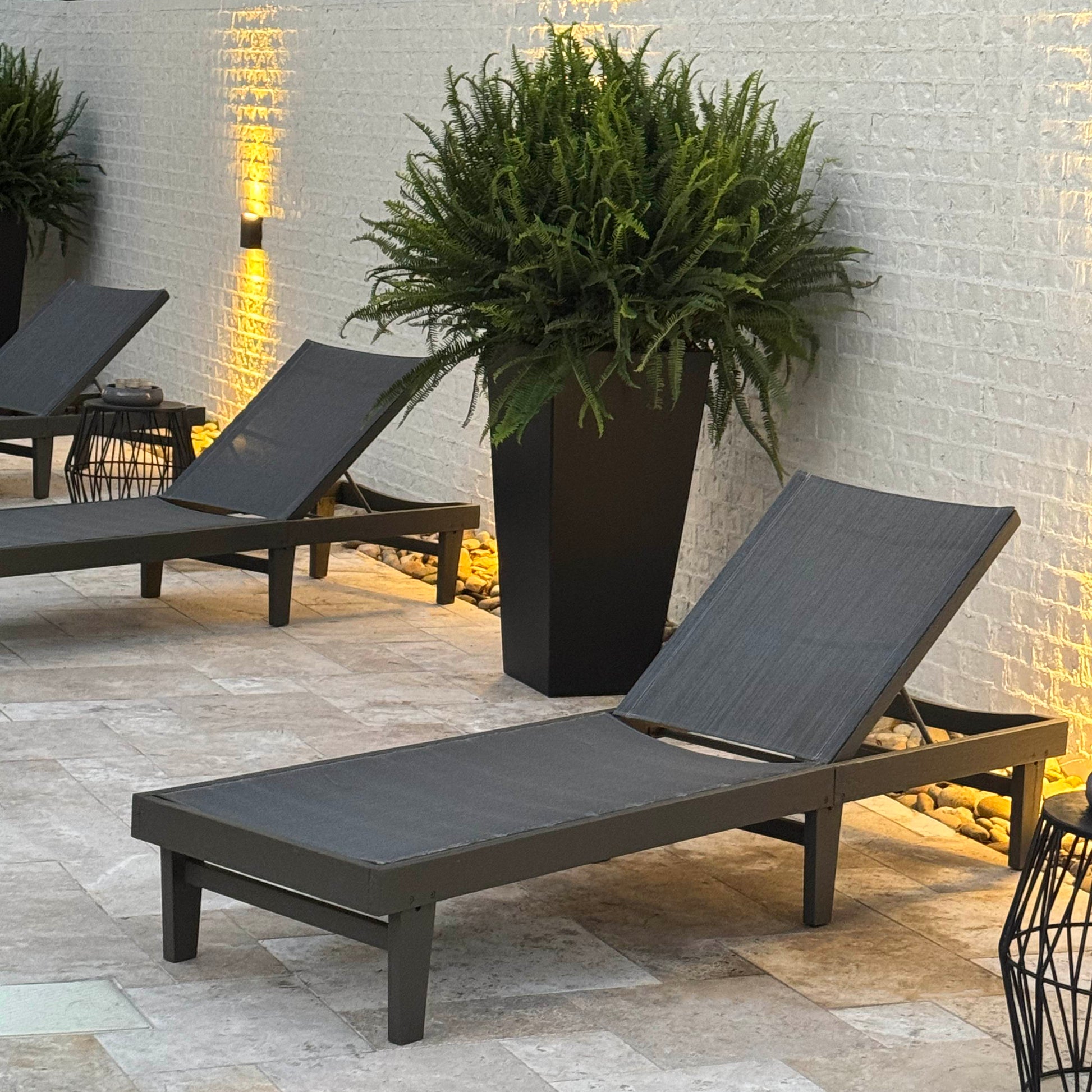 Tall black planter with lush fern beside modern patio loungers and uplighting.