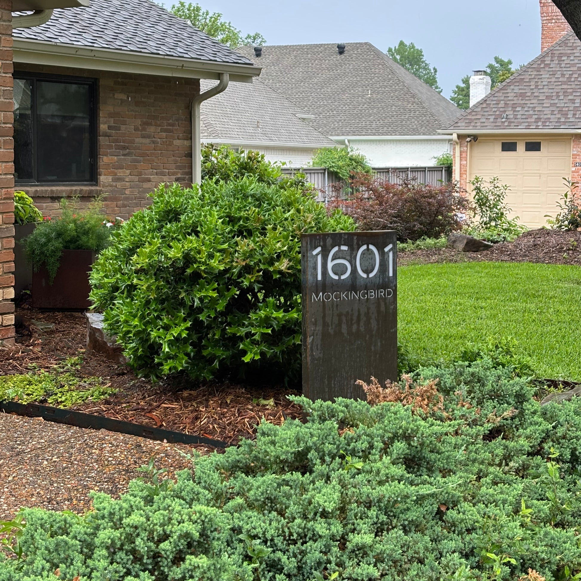 Corten steel address sign in landscaped front yard with modern house numbers.