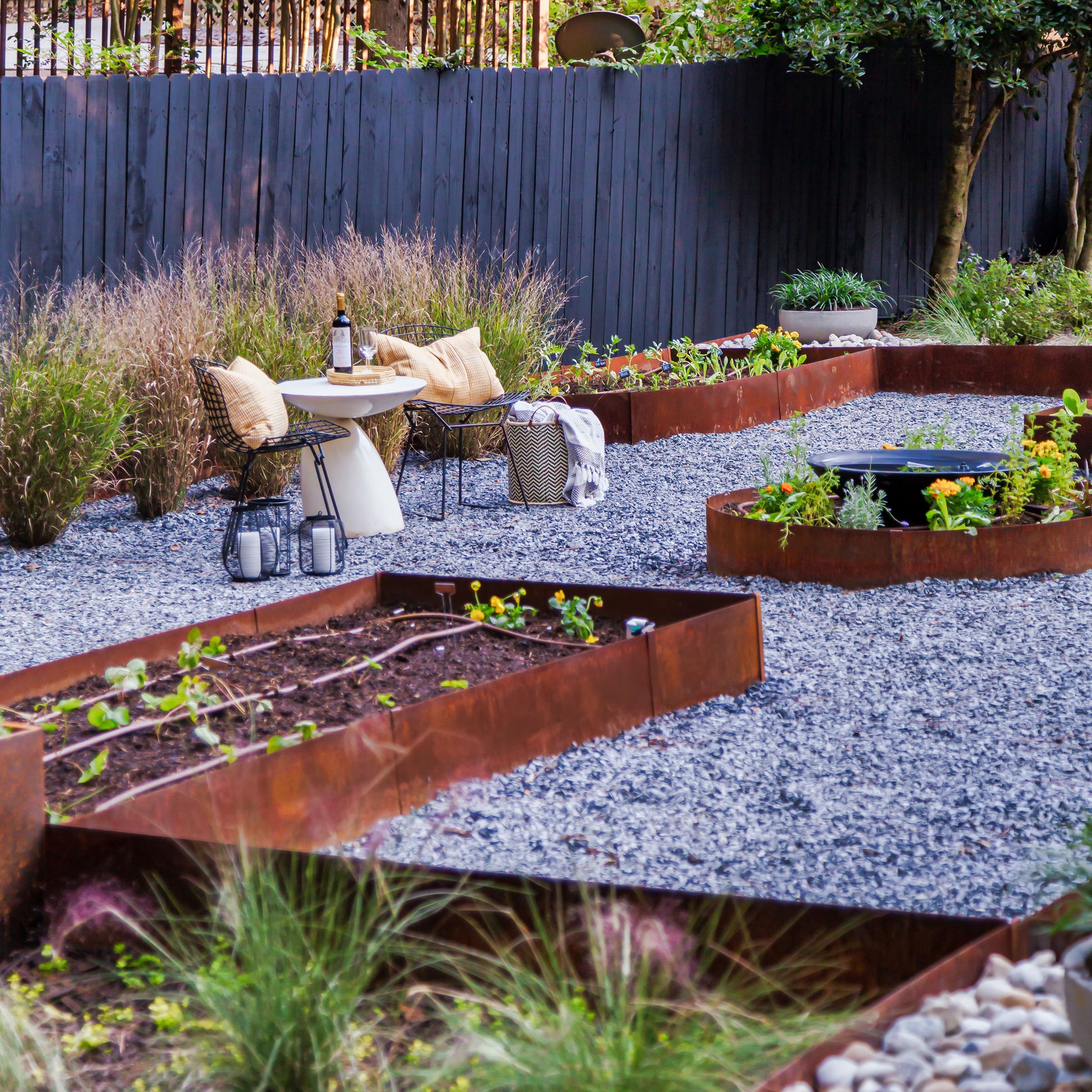 gravel area with raised beds that are constructed with corten steel metal landscape edging that has developed a protective patina. The raised beds vary in size and shape and contain different species of flowers. Three is a small sitting area that consist of a table and two chairs. 